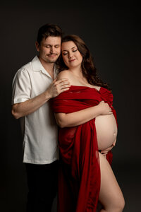 Expecting couple embracing during a studio maternity photoshoot with mother in a red gown photographed by Kaitlyn Dawn Photography.