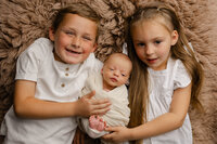 Big siblings kissing their newborn baby on a dark brown backdrop. Photographed by Inland Empire newborn photographer Kaitlyn Dawn Photography.