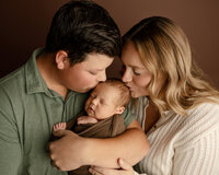 Mother and father kissing their newborn baby’s head while holding their sleeping baby during an emotional studio newborn family photography session in Yucaipa by Kaitlyn Dawn Photography.