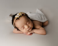 Sleeping newborn baby girl posed with hands under chin and wearing a floral headband on a soft cream backdrop during a studio newborn photography session by Yucaipa newborn photographer Kaitlyn Dawn Photography.
