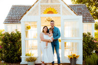 a couple smiles with their baby girl in front of a greenhouse in leonard texas.