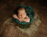 Sleeping newborn baby boy posed in a green bucket prop on a rustic wooden backdrop during a studio newborn photography session by Kaitlyn Dawn Photography.
