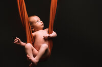 A naked baby lies on an orange fabric against a black backdrop. The orange fabric cradles the baby like a swing.