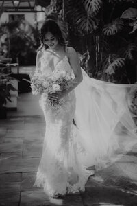 Mariée souriante tenant son bouquet, robe de dentelle à traîne fluide, devant un décor de plantes tropicales à Aquatopia, Ottawa, portrait en noir et blanc