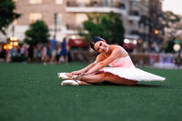 A ballerina sits in the grass, reaching for her toes and smiling brightly. A downtown scene is bokehed behind her.