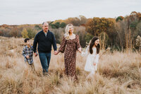 Family of four holds hands and walks in field during fall family photography session