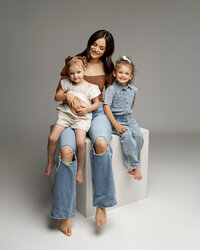 Mother sitting in studio holding her two daughters on her lap, relaxed and joyful family moment captured by Kaitlyn Dawn Photography, professional family photography studio.