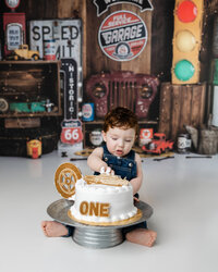 Baby boy in denim overalls digging into a white cake topped with a gold “ONE” and tire-themed decoration, set in a vintage garage backdrop with Route 66 details. Photograph by Yucaipa cake smash photographer Kaitlyn Dawn Photography.