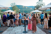 A crowd of well dressed guests chats and laughs under string lights on a patio, with mountain views and decorative umbrellas.