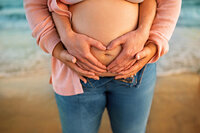 Close up detail of expecting parents forming a heart with hands over pregnant belly at Atlantic beach maternity session