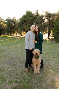 Couple embracing beside their dog in a grassy outdoor setting during golden hour, candid lifestyle portrait in Yucaipa by Kaitlyn Dawn Photography.