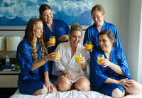 Bride and bridesmaids in matching robes laughing and clinking mimosas during a joyful wedding-morning getting-ready moment, captured by a Colorado wedding photographer.