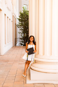 Senior girl holding cap and wearing cords while leaning against large pillar outside her college on the Belmont campus