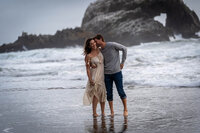 man whispering to a woman in a dress as they lovingly walk down the beach without shoes as they wade in the water