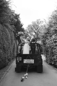 Black and white wedding photograph of newlyweds standing in the back of a Land Rover at Avington Park, Hampshire, driving along a tree-lined country lane with a handmade “Just Married” sign and tin cans trailing behind.