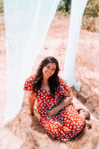 Singer-songwriter seated outdoors in a red floral dress beneath flowing white fabric