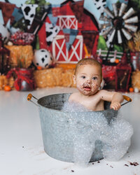 Baby with chocolate on their face enjoying a bubble bath in a metal tub after cake smash. Photograph by Yucaipa cake smash photographer Kaitlyn Dawn Photography.