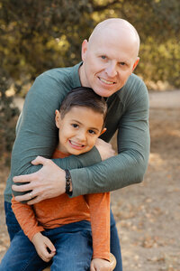 Father hugging his young son outdoors in warm natural light, candid family portrait by Kaitlyn Dawn Photography, lifestyle family photographer specializing in authentic moments.