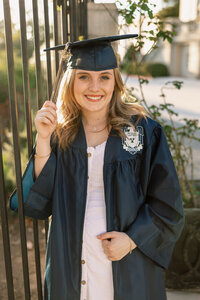 High school graduate smiling while adjusting her cap and gown during sunset, senior graduation portrait by Kaitlyn Dawn Photography.