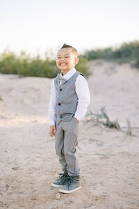 Young boy from the Collins family smiling brightly in the Las Vegas desert, wearing a gray vest and bow tie during a golden hour family photography session.