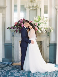 bride leaning into groom as he holds her in front of a floral fireplace in a soft romantic setting