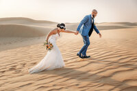 Wedding portrait of a bride and groom walking across desert dunes in Dubai. Dubai desert elopement. Intimate wedding planned by Lovely & Planned.