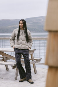 Model in a taupe down puffer and black corduroy trousers beside a picnic table on the dock