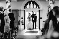 Black and white photo of bride and her father walking down church asile