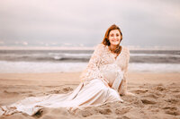 A redheaded pregnant woman sits on a beach. The water and horizon are behind her. The sky is blue with subtle shades of pink.