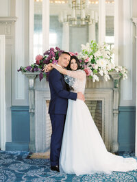 bride and groom embracing in front of a fireplace adorned with romantic floral arrangements in soft pastel tones