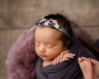 Close-up portrait of a sleeping newborn baby girl wearing a floral headband and wrapped in a purple swaddle during a studio newborn photography session by Kaitlyn Dawn Photography in Yucaipa.