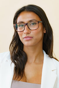 Young woman with long hair, a white jacket, and glasses smiling for a headshot photo