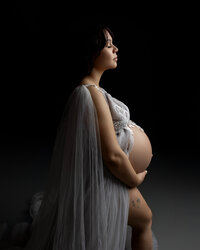 Fine art maternity photography portrait of pregnant woman in profile wearing a sheer flowing white maternity gown with soft studio lighting emphasizing her baby bump by Yucaipa best maternity photographer Kaitlyn Dawn Photography.