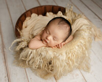 Sleeping newborn baby posed in a wooden bowl with a soft cream flokati blanket during a studio session with newborn photographer Kaitlyn Dawn Photography.