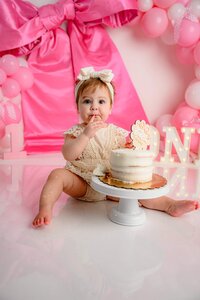 little one year old girl dressed in cream sits in a pink bow cake smash set up taking a bite of her cake