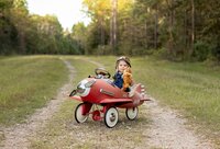 Professional photography by Nature's Child Photography: Baby wearing an aviator hat and overalls sitting in a red vintage pedal airplane and holding a stuffed teddy bear on a wooded trail during an outdoor child portrait session in St. Johns.