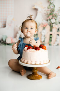 One-year-old sitting with a strawberry-topped birthday cake in a light, airy studio setup. Photographed by Yucaipa baby photographer Kaitlyn Dawn Photography.
