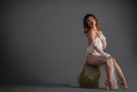 Woman in a pale robe seated on a geometric block against a dark studio background, one leg extended.