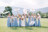 Bridesmaids in light blue dresses with white parasols on the lawn at King Family Vineyard wedding in Charlottesville, VA — floral design by Flowergirl Cville | Photo by Paige Ryan Photography