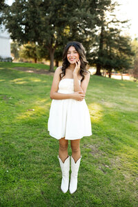 High school senior girl standing on a grassy lawn wearing a white dress and boots, natural outdoor senior portrait by Kaitlyn Dawn Photography.