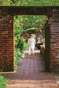 Young girl in white lace dress walking barefoot through a brick garden walkway framed by an arch