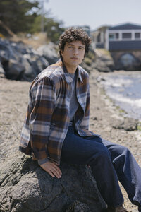 Brown and blue plaid overshirt over a grey knit with denim, seated on the rocks