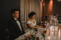Mariés souriants à la table d'honneur, robe bustier blanche et smoking noir, décor floral et chandelles, ambiance chaleureuse à la Gare Viger, Montréal