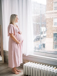 Pregnant mom wearing a linen pink dress standing in a studio looking outside at the falling snow