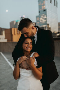 Groom tenderly kisses the bride on the head during a rooftop sunset, creating a romantic and memorable moment