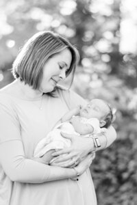 A black and white photo of a mom cradling a newborn baby in her arms while the light filters through the tress in the background