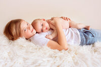 Older sister lying on cream blanket holding newborn baby during studio portrait session