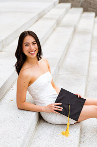 Classic senior graduation photo on the stairs at Vanderbilt University. Senior wearing white dress smiling at camera with cap in her hand.