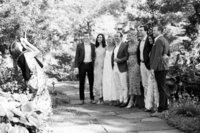 A group of eight friends poses for a photo in a garden setting, while a woman in a floral dress captures the moment with her phone.