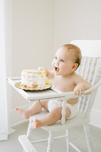 Baby boy in white high chair smashes first birthday cake  during baby photography session in Raleigh NC
